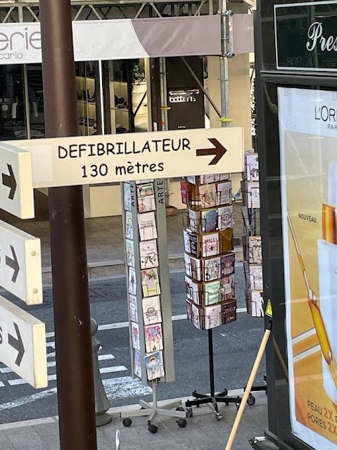A street sign points 130 meters to a defibrillator, while a nearby stand showcases postcards enticing you to travel the French Riviera in the off season. In the backdrop, a crosswalk and a cosmetics ad complete this charming urban scene.