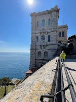 A tall, historic building stands near the edge of a scenic coastal pathway along the French Riviera, enjoying clear blue skies. The sun shines brightly, casting playful shadows as the sea remains calm and expansive. A person in a yellow jacket strolls down the railing-lined path, embracing off-season tranquility.