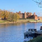 A large historic building with many chimneys sits across the river along the Thames Path, framed by autumn trees and a blue sky. Several boats are moored in the foreground along the riverbank.
